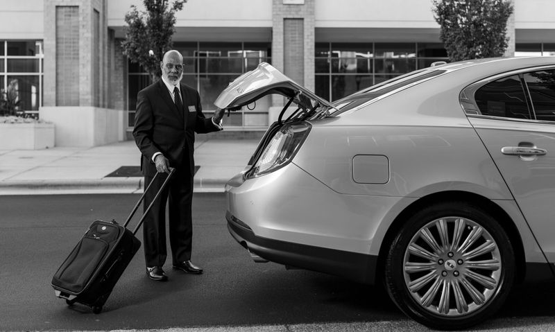 A chauffeur with a rolling suitcase at the open trunk of a sedan